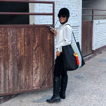 Load image into Gallery viewer, Person in equestrian attire with a helmet and boots, holding a bag, standing next to a wooden gate.