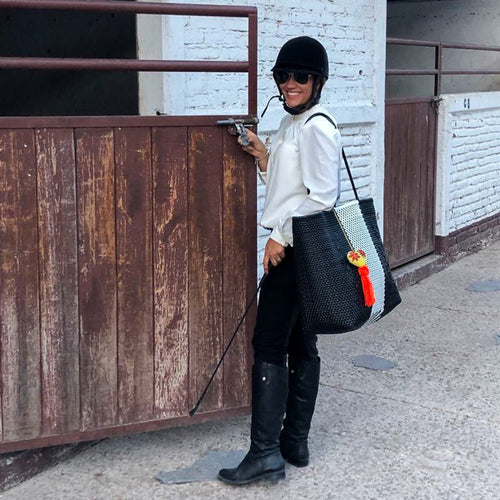 Person in equestrian attire with a helmet and boots, holding a bag, standing next to a wooden gate.